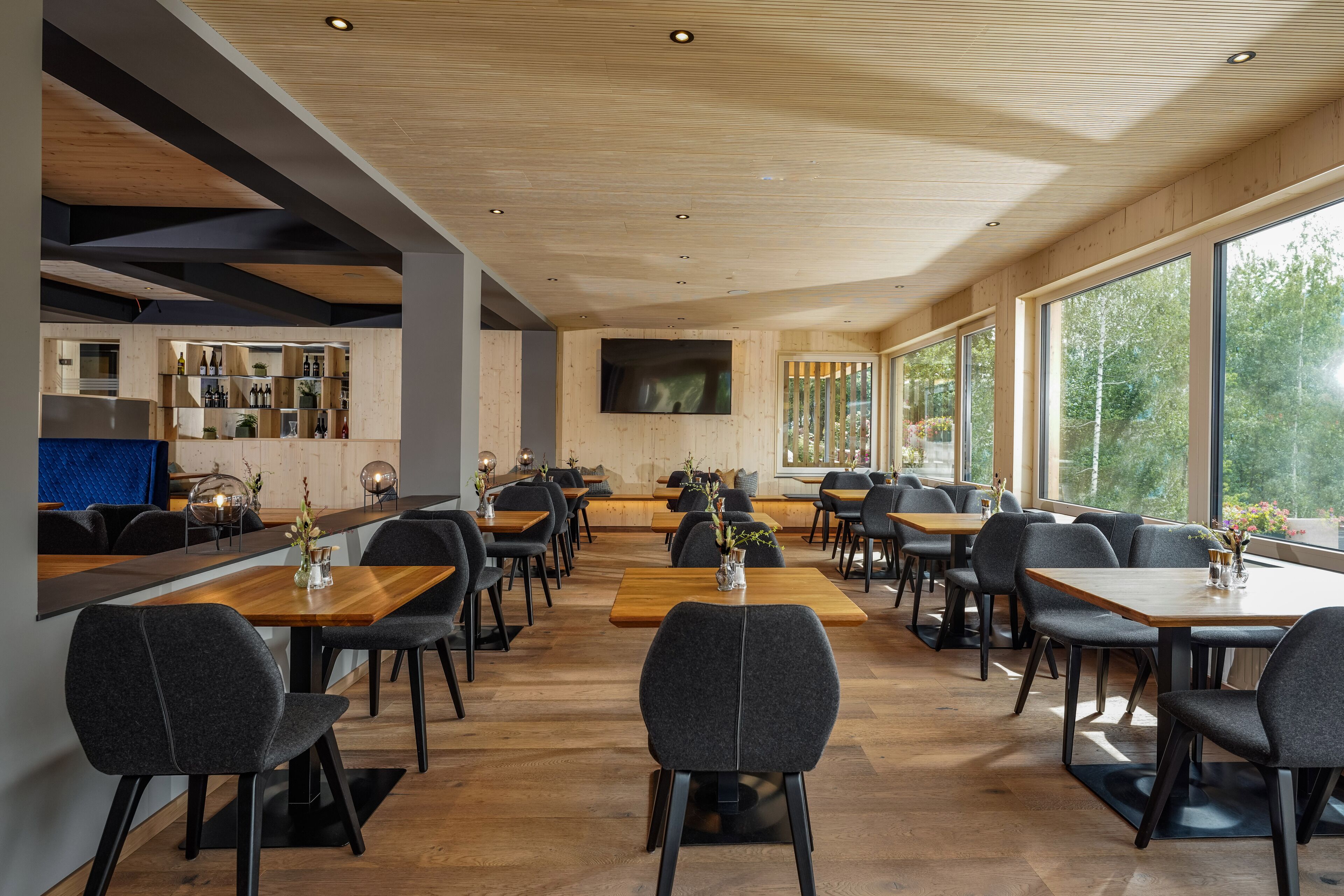 Un restaurant moderne avec des planchers en bois, des chaises noires et de grandes fenêtres donnant sur la verdure.