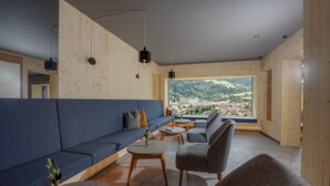 A modern interior with a large window offering a scenic view, a blue sofa, grey chairs, and wooden tables. - Ferienalm Panorama Hotel & Apartments (Schladming)