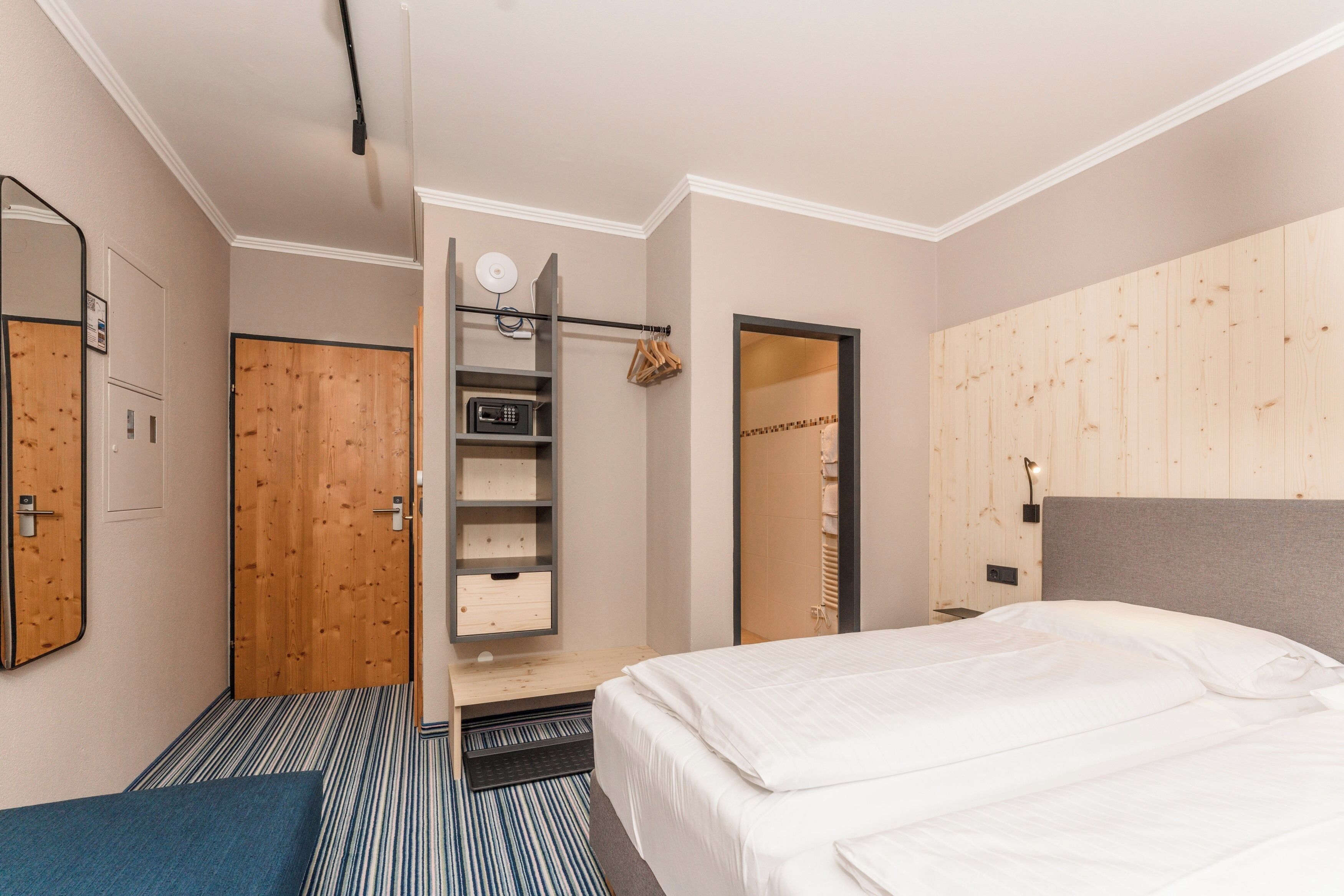 A hotel room with a bed, a wardrobe, a mirror, a shelf with storage, and a door leading to another room.