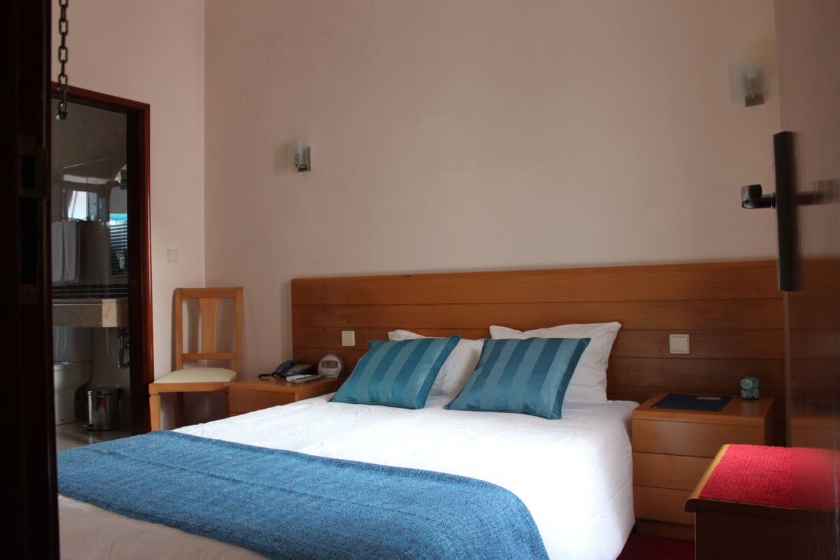 A hotel room with a wooden headboard, a large bed with blue and white bedding, a wooden nightstand with a phone, and a red bench.
