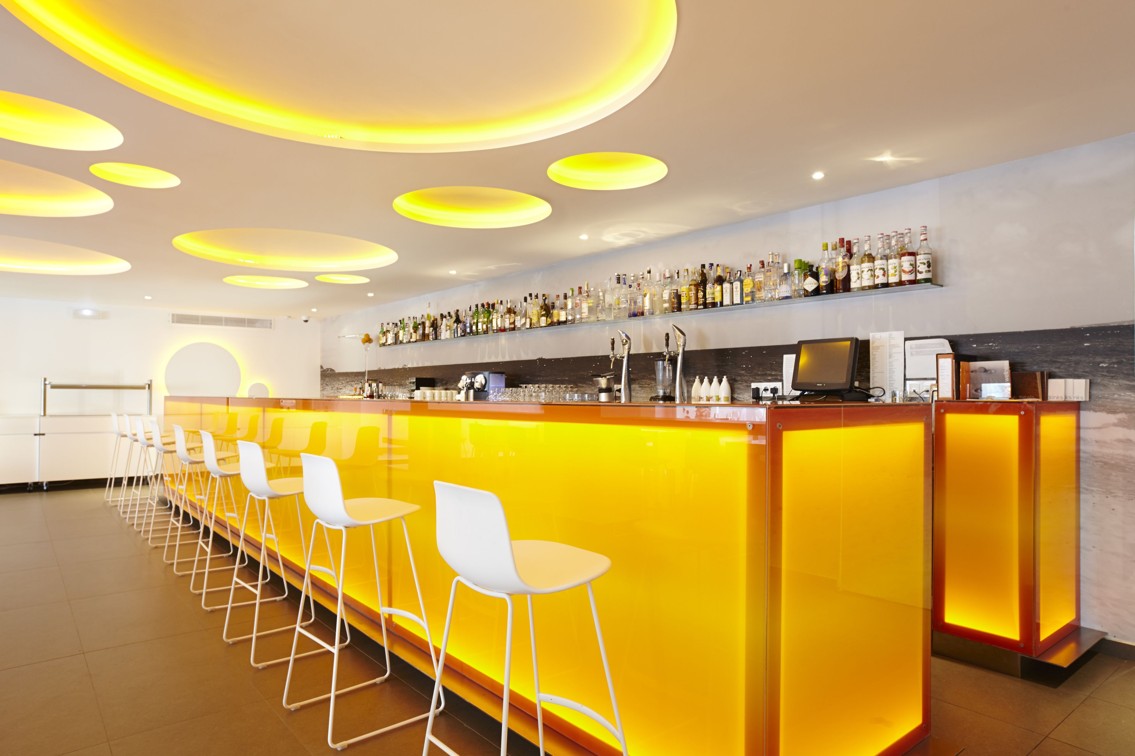 A bar area with a long counter, white bar stools, and a shelf with bottles.