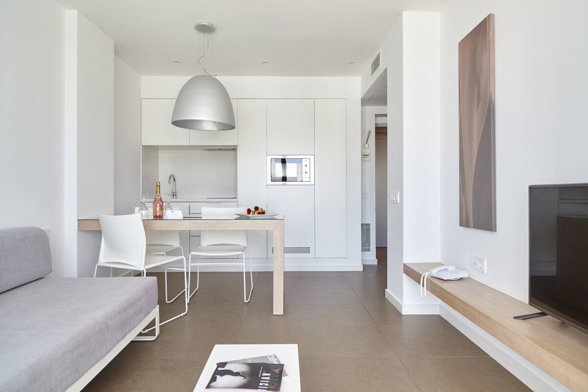 A modern, minimalist living room with a grey sofa, a dining table with chairs, a pendant light, and a flat-screen TV on a wooden shelf.