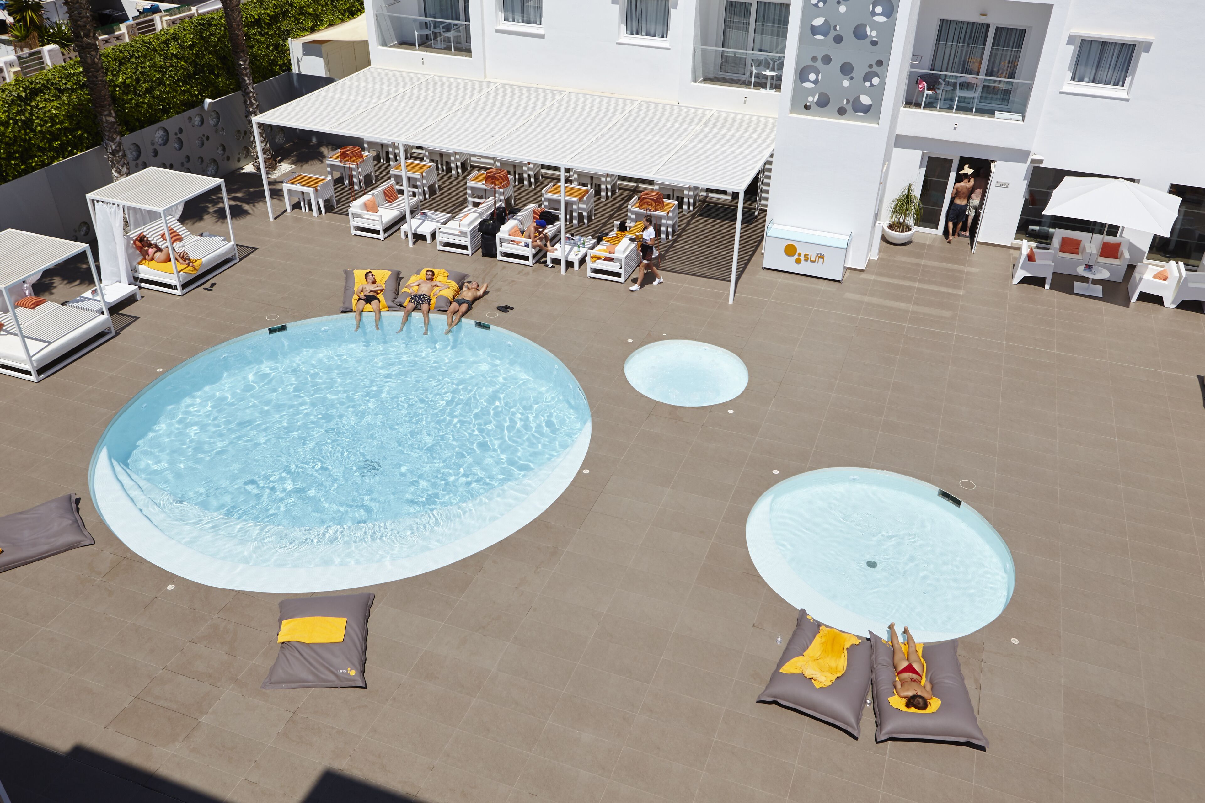 Un espace piscine sur le toit, comprenant de nombreux chaises longues et parasols, une grande piscine centrale et une piscine plus petite attenante.
