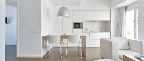 A modern, minimalist living room with a dining area, a kitchen, and a large window providing natural light.
