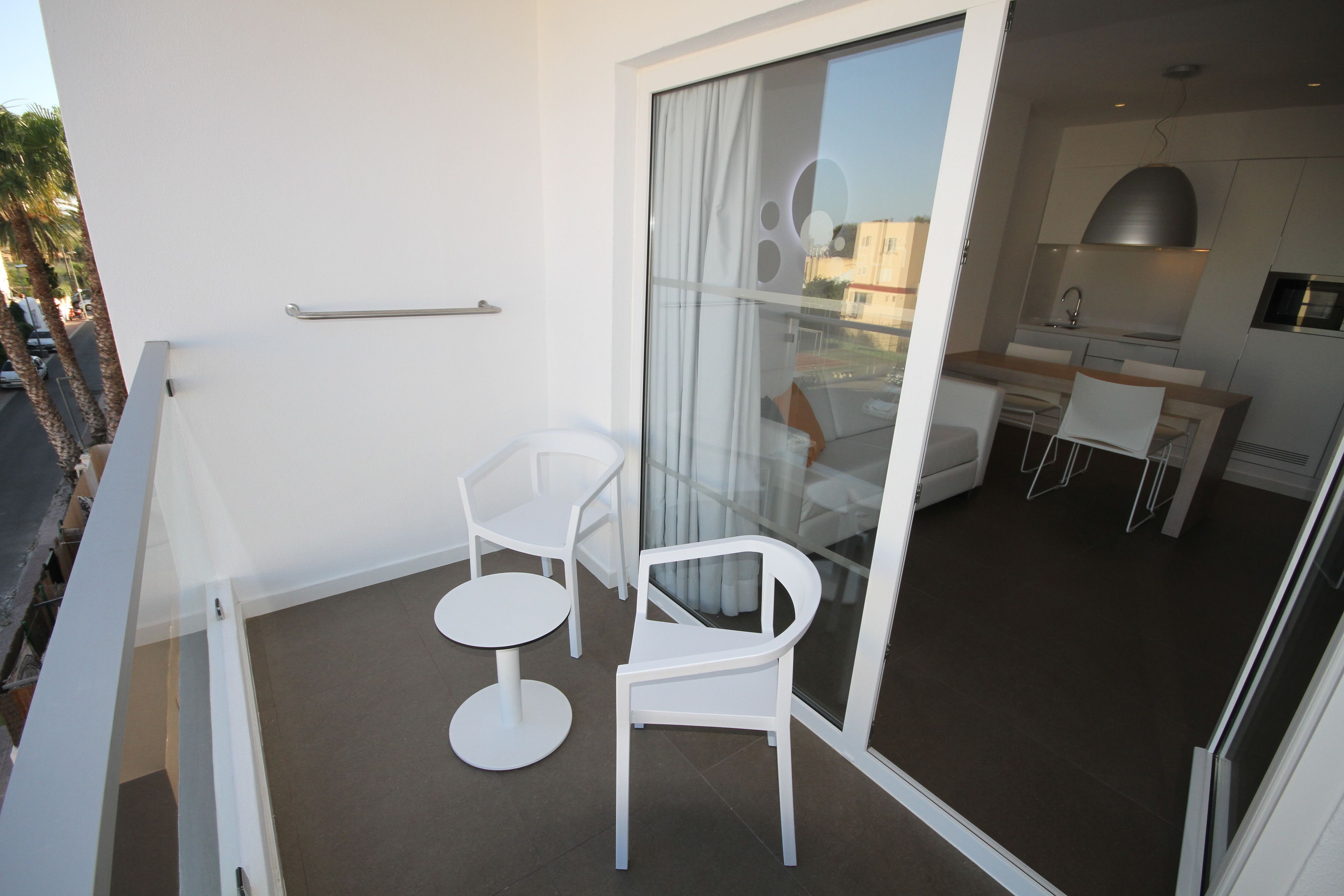 Un balcón moderno con sillas blancas y una mesita, una puerta corrediza de vidrio que da a una zona de cocina con una mesa y sillas para comer.