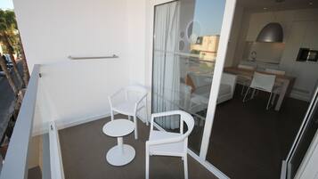 Un balcón moderno con sillas blancas y una mesita, una puerta corrediza de vidrio que da a una zona de cocina con una mesa y sillas para comer.