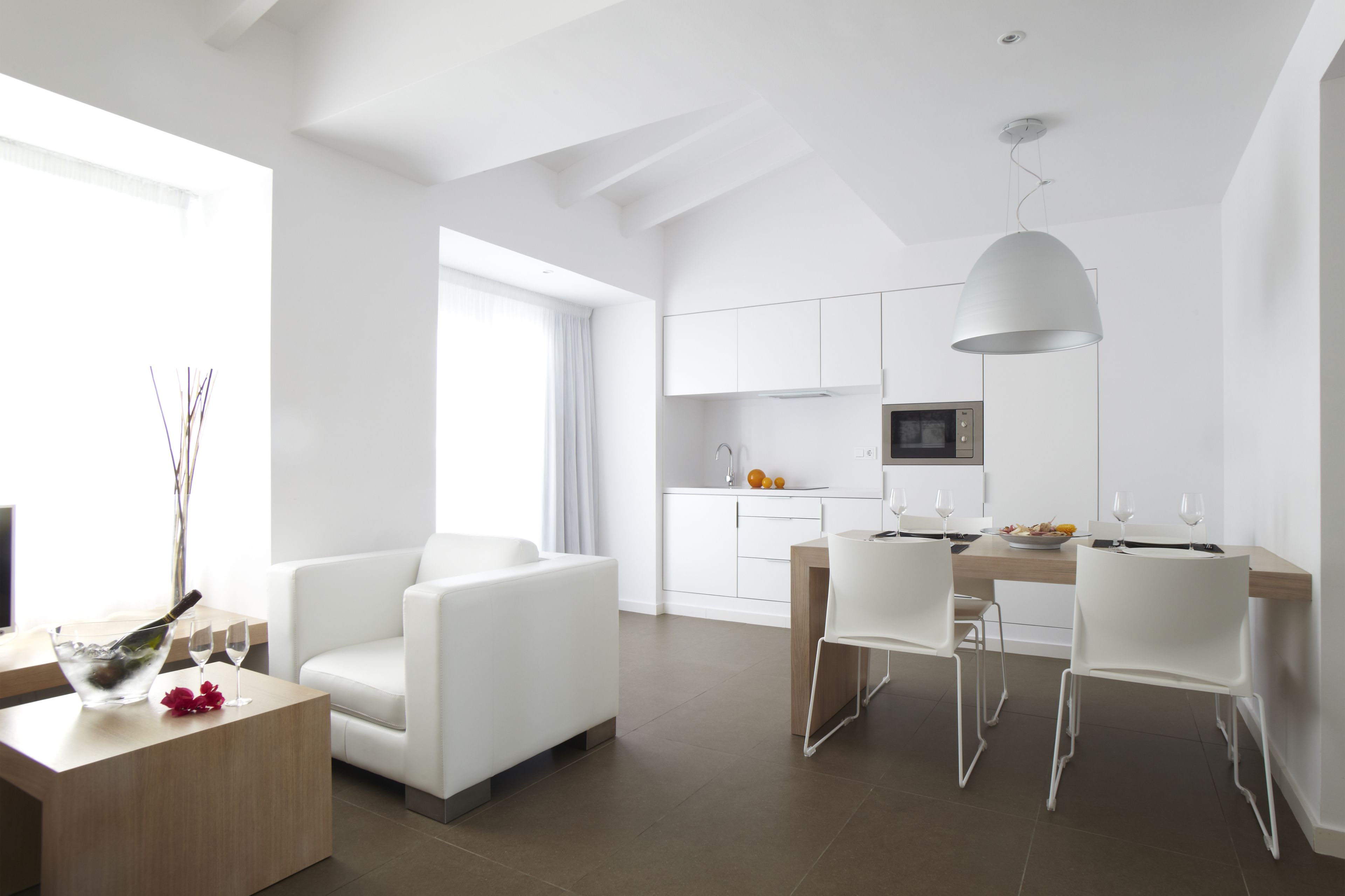 Un área de estar moderna y bien iluminada con una mesa de comedor, sillas blancas, un sofá y una cocina al fondo.