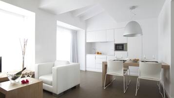 Un área de estar moderna y bien iluminada con una mesa de comedor, sillas blancas, un sofá y una cocina al fondo.