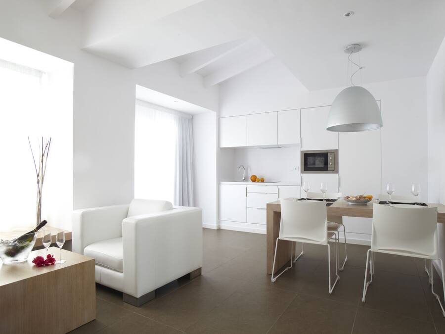 Un área de estar moderna y bien iluminada con una mesa de comedor, sillas blancas, un sofá y una cocina al fondo.