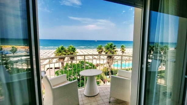 A balcony with a view of the beach, palm trees, and a pool. - Hotel Ca' Bianca (Riccione)