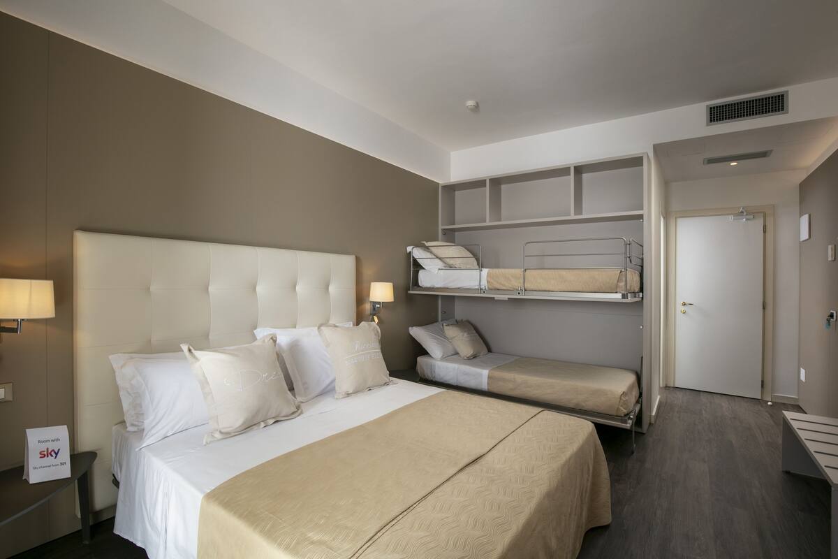 A hotel room with a large bed, a smaller bed, and a bunk bed. There is a desk with a lamp, a door, and a sign indicating room availability.