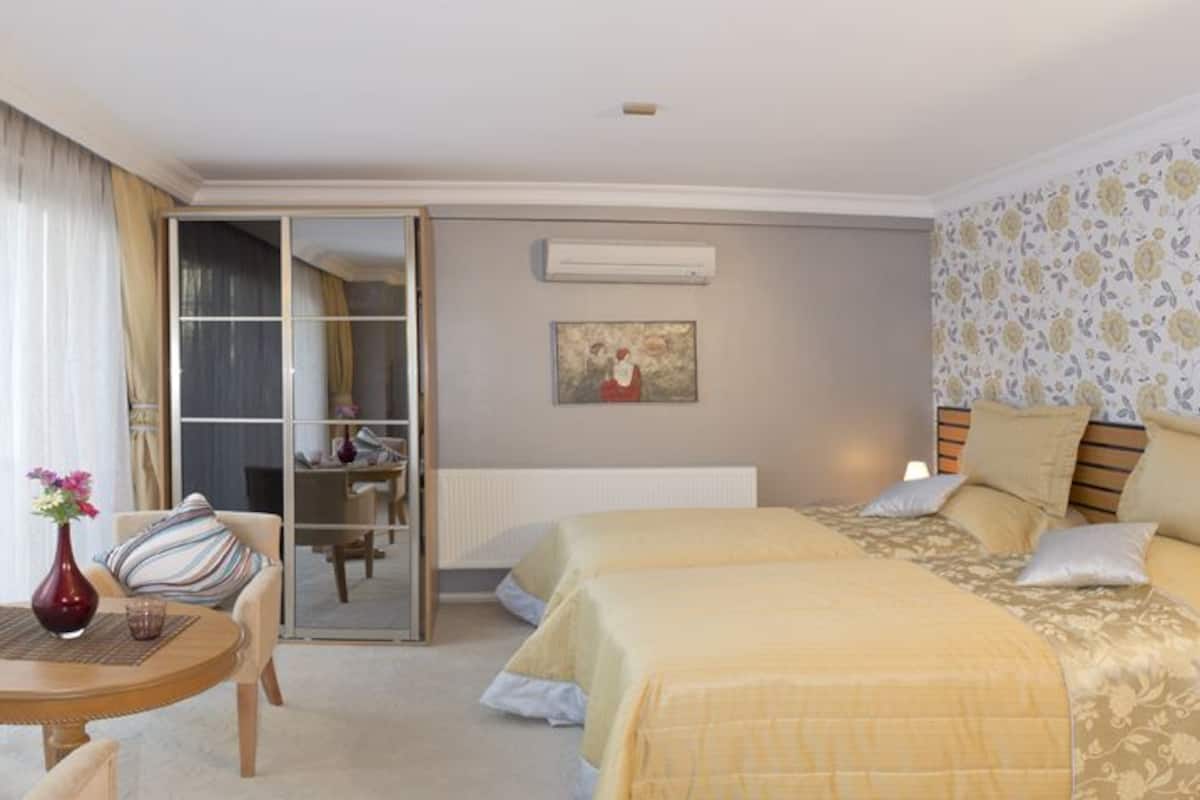 A hotel room with two beds, a small table with a vase, a chair, and a view of the outside through a sliding door.