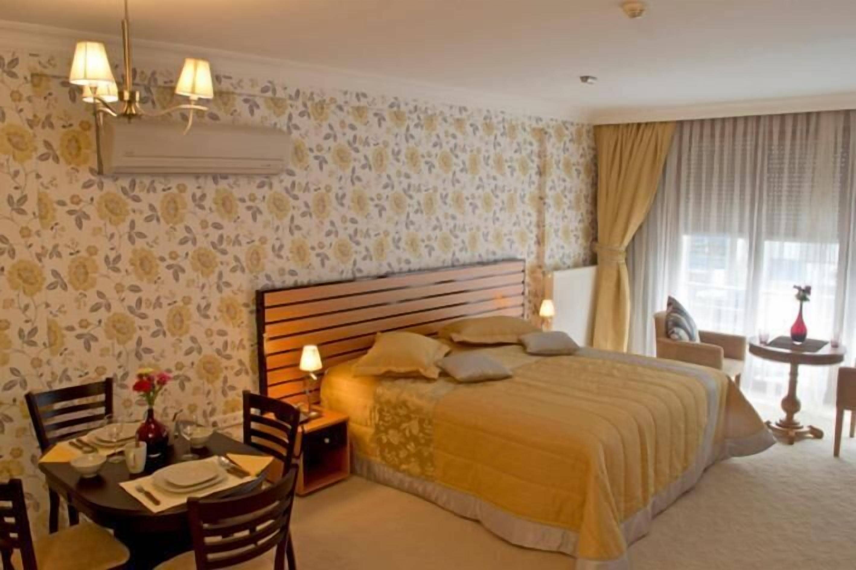 A hotel room with a large bed, a bedside table with a lamp, a small round table with a vase, and a chair.