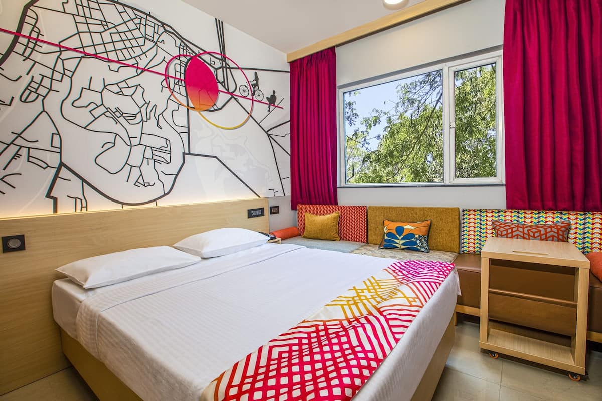 A hotel room with a large bed, a map mural on the wall, a window with red curtains, and a wooden nightstand.