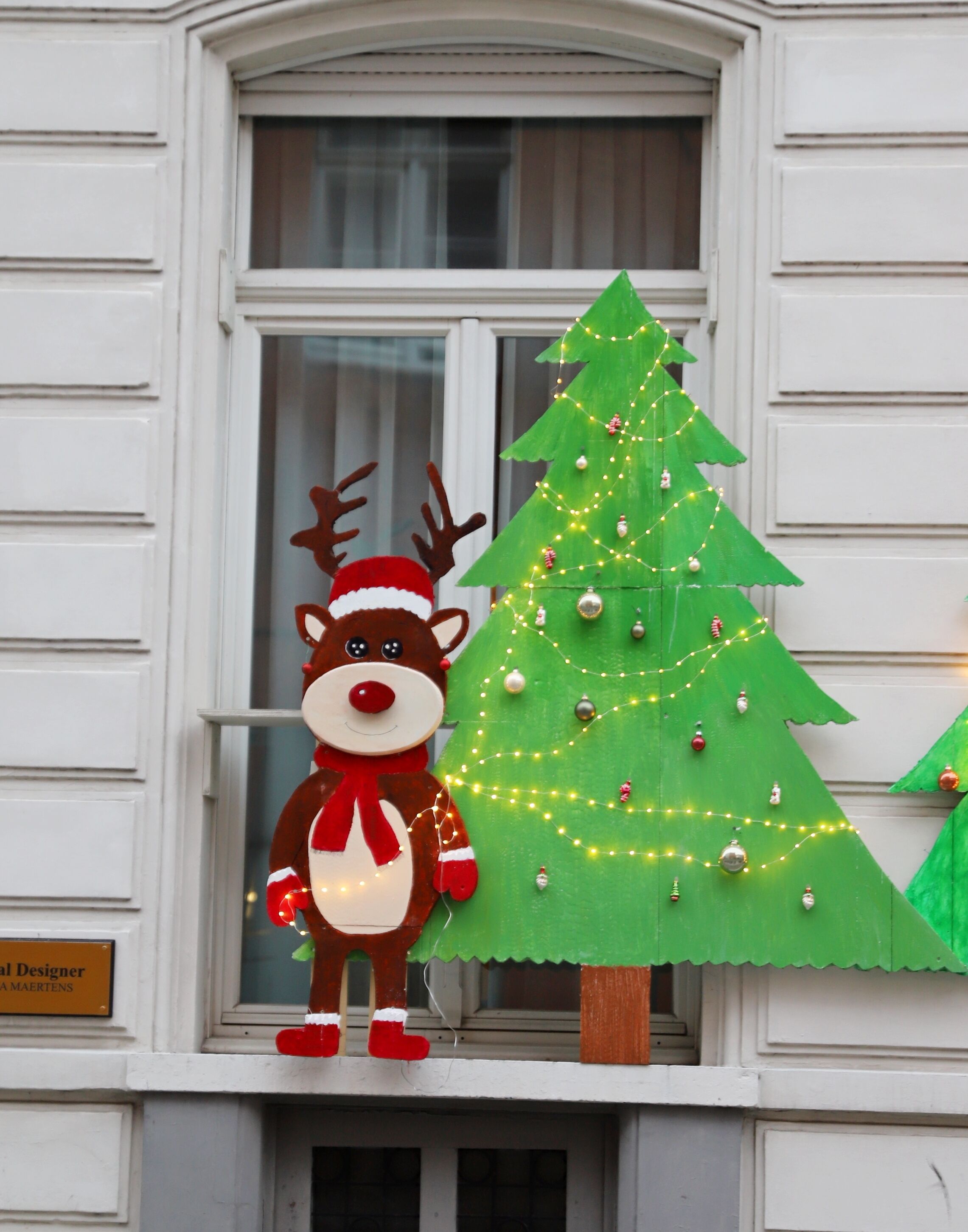 Eine Gebäudefassade mit Fenster, Rentier-Dekoration und einem Weihnachtsbaum mit Lichterketten.