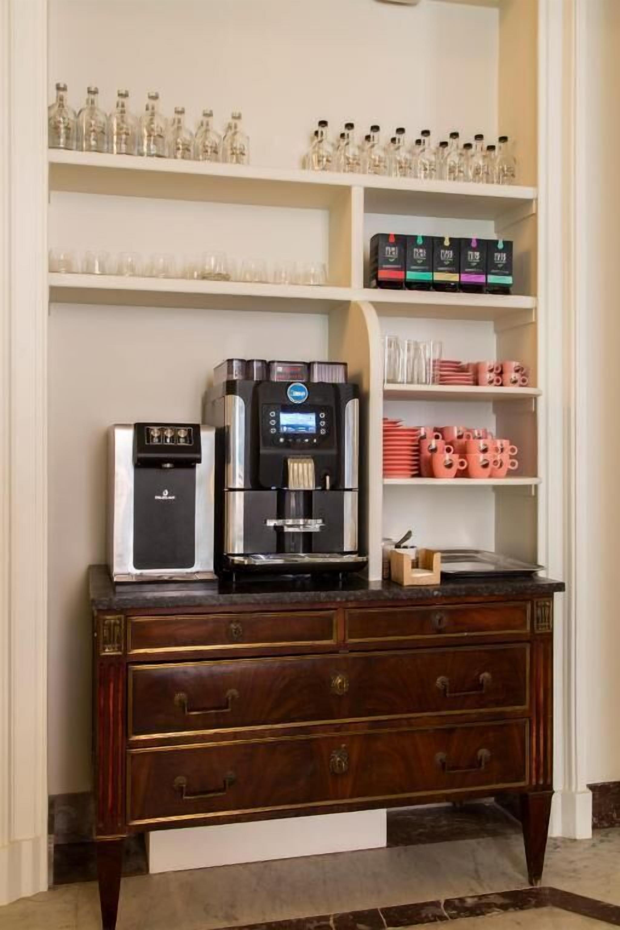 A coffee station with a coffee machine, a water dispenser, and a variety of mugs and cups.