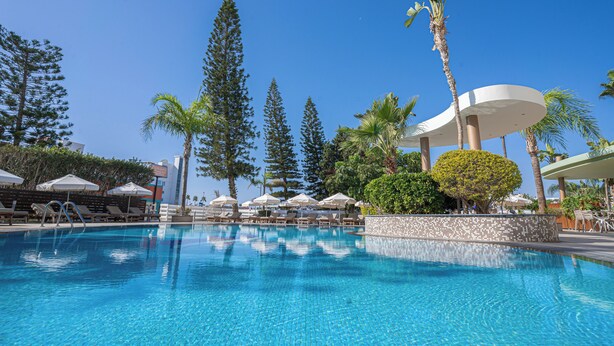 A swimming pool with lounge chairs and umbrellas, surrounded by tall trees and a palm tree. - Stamatia Hotel (Ayia Napa)