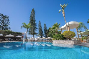 Une piscine entourée de chaises longues et de parasols, bordée de grands arbres et d’un palmier.
