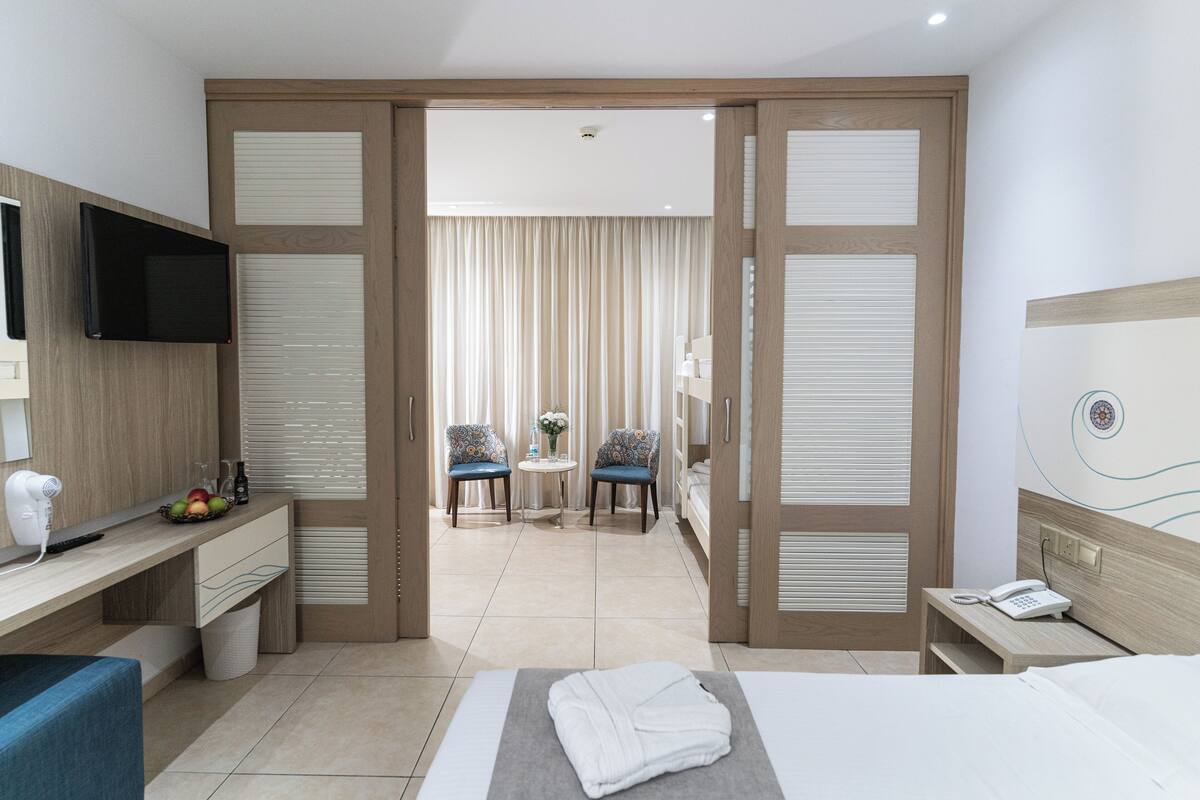 Ein Hotelzimmer mit Bett, Nachttisch, Fernseher und Schreibtisch mit Stuhl. Im Hintergrund steht ein kleiner Tisch mit Stühlen.