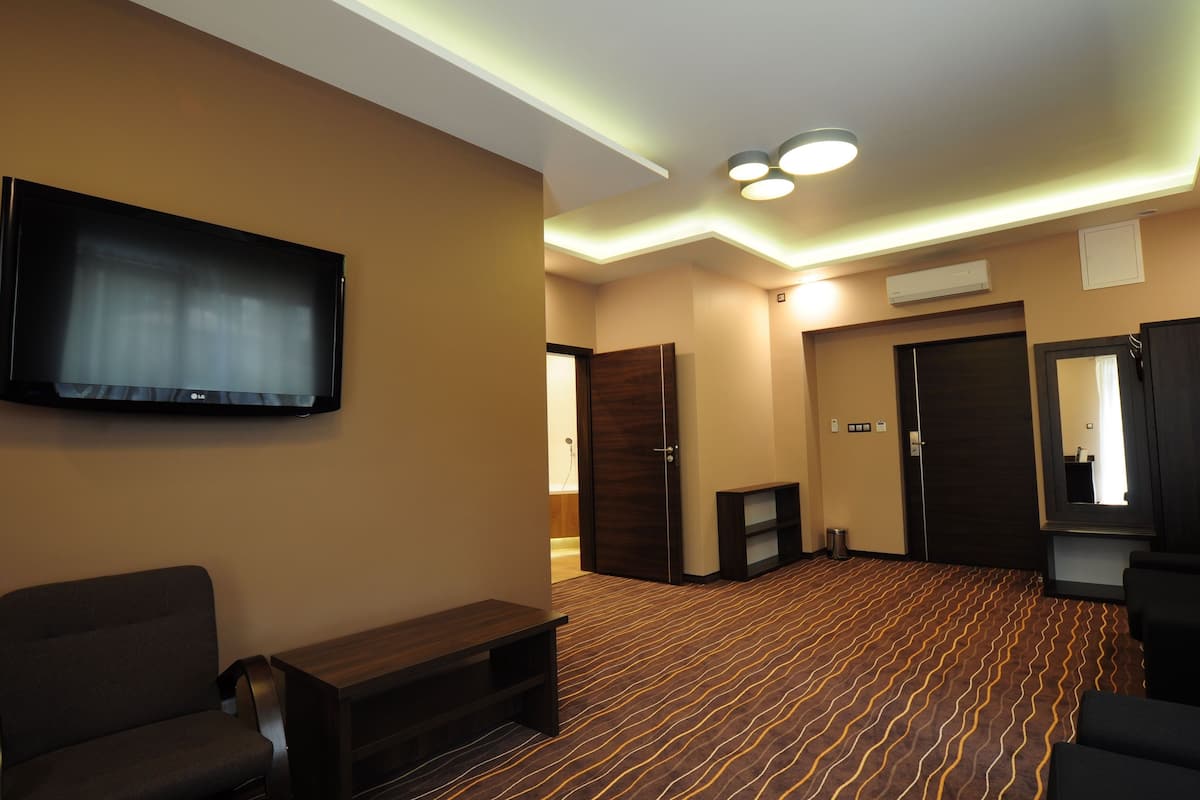 Una habitación de hotel con televisión de pantalla plana, un banco de madera oscura, una alfombra estampada y una puerta que da a otra habitación.