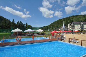A swimming pool with a wooden deck, surrounded by beach chairs and umbrellas, with a hotel and forested hills in the background. - Hotel Zimnik Luksus Natury (Lipowa)