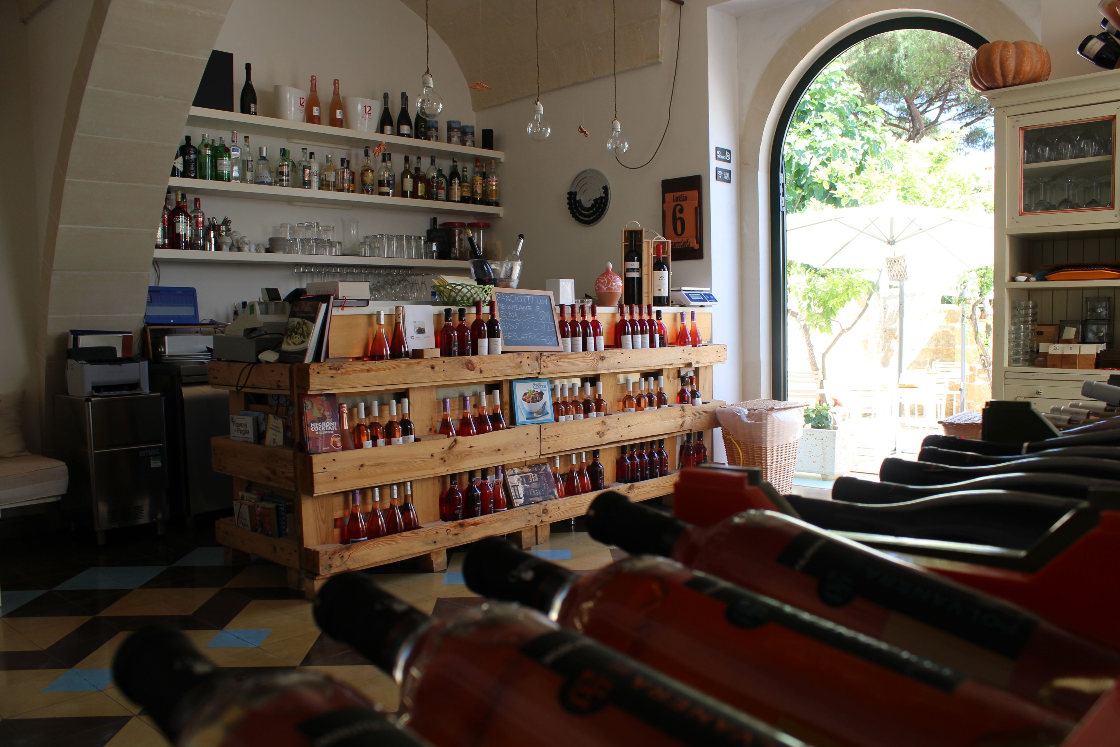 A bar area with shelves of bottles and glasses, a chalkboard menu, and a view through an arched doorway.