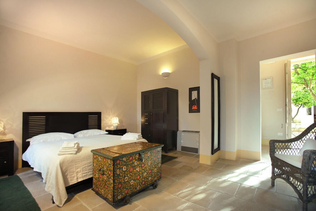 Un dormitorio con cama, mesitas de noche, cómoda, banco y puerta que da a un balcón.