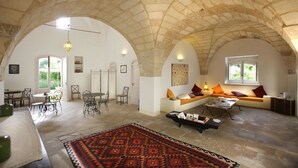 A spacious room with a high arched ceiling, a dining area with a table and chairs, a seating area with cushions, and a patterned rug on the floor. - Palazzo Guglielmo Albergo Diffuso (Ortelle)