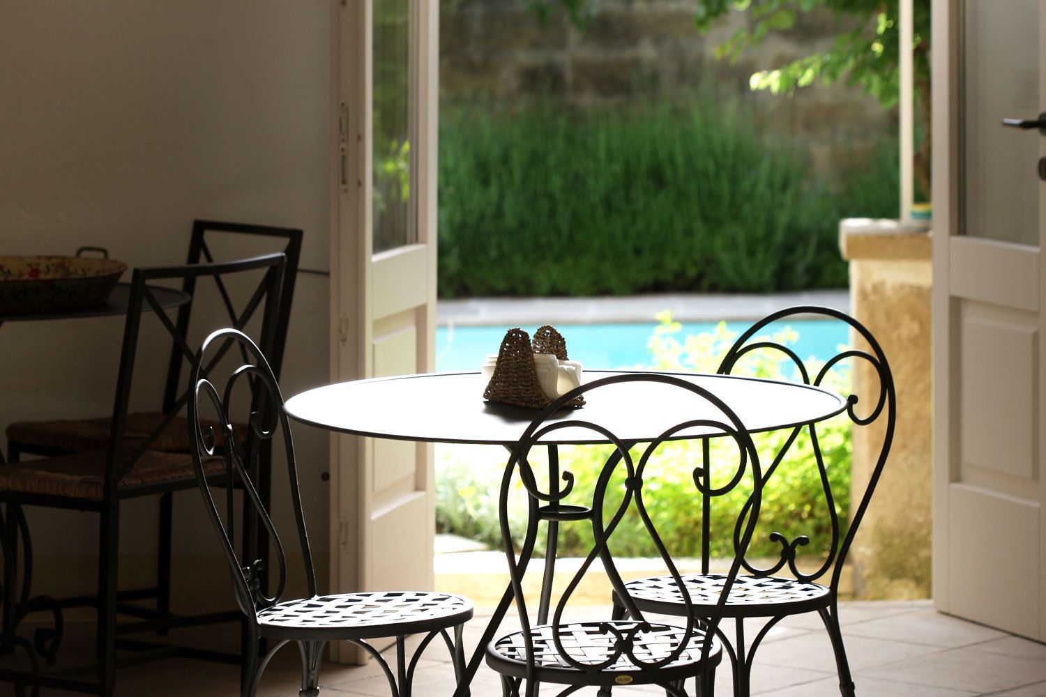 A dining area with a round table and two chairs, a small decorative item on the table, and a view of a swimming pool and greenery outside.