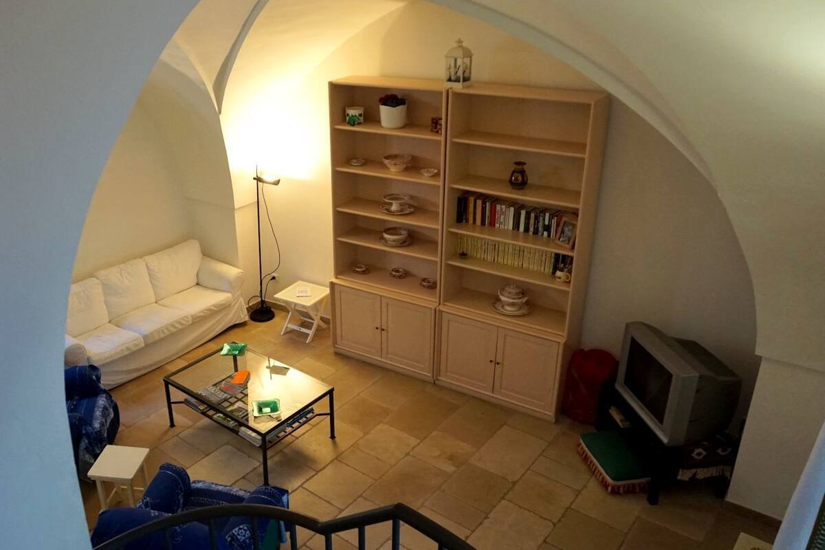 Una sala de estar acogedora con un sofá blanco, una mesita de centro y un mueble para televisor. Hay una estantería llena de libros y objetos decorativos.