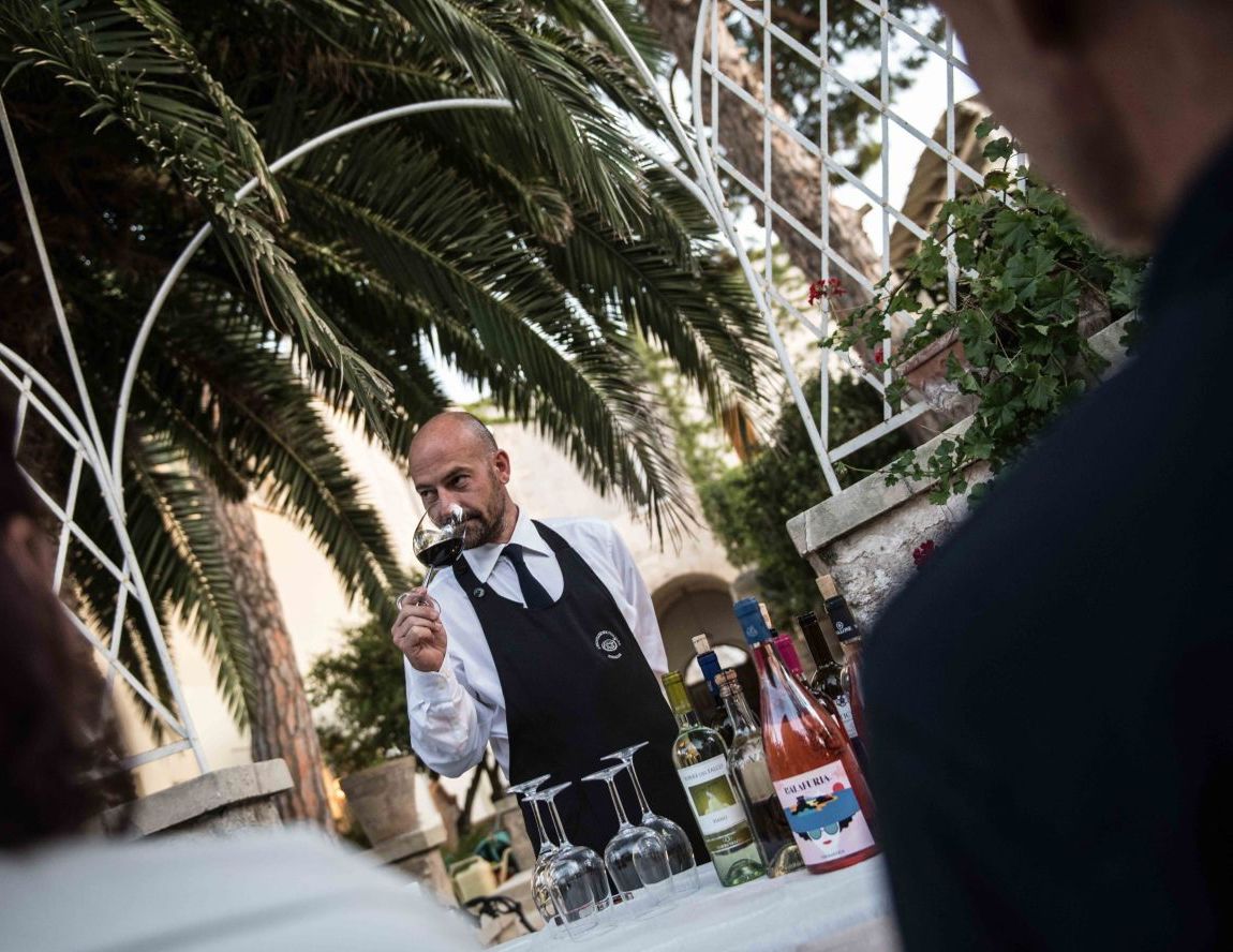 A person serving wine at an outdoor table with a tropical backdrop.
