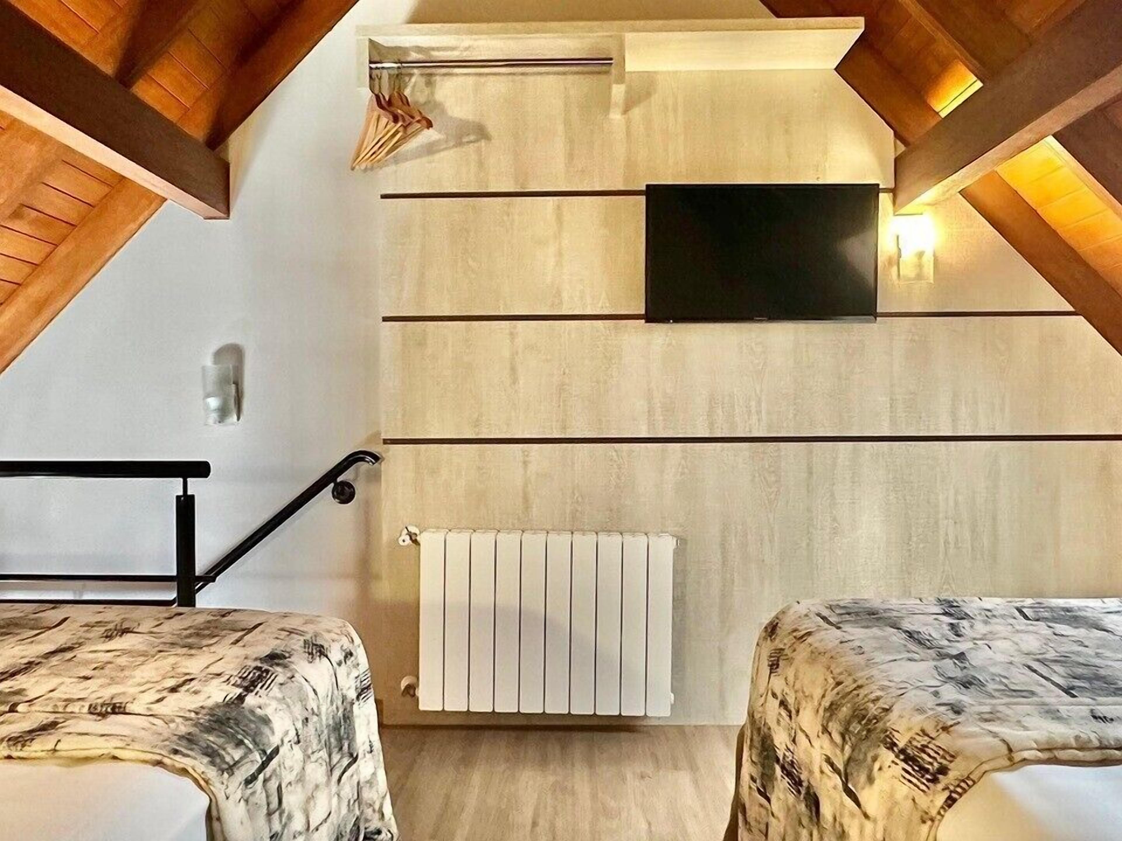 Quarto com teto inclinado, televisão montada na parede, radiador e cama com cobertura estampada.