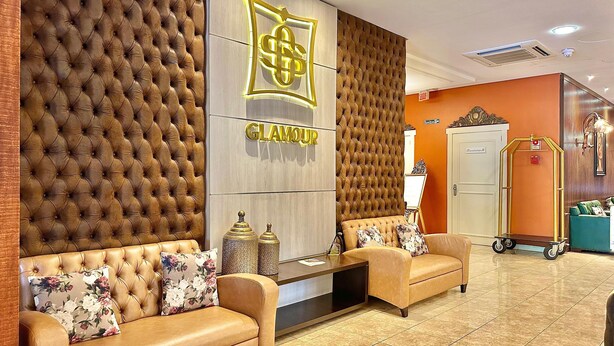 A reception area with a tufted wall, a 'GLAMOUR' sign, two sofas with floral cushions, a small table, and a wardrobe. - Hotel Glamour da Serra (Gramado)