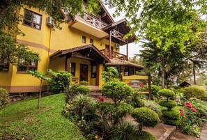 A yellow building with a balcony, surrounded by well-maintained gardens and trees. - Pousada das Papoulas (Gramado)