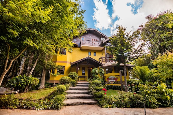 A yellow building with a balcony, surrounded by lush greenery and a stone staircase leading to the entrance. - Pousada das Papoulas (Gramado)