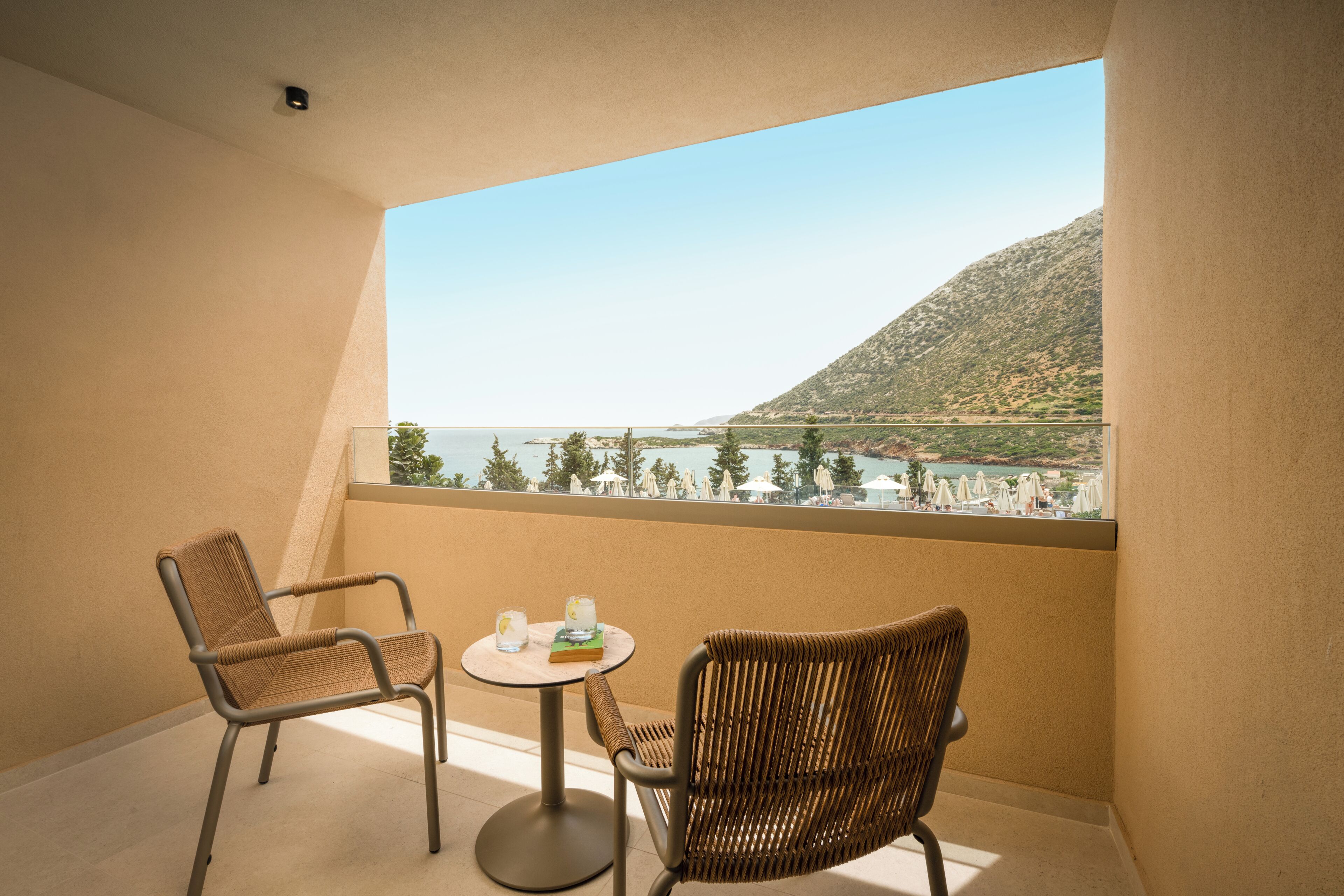 A balcony with two wicker chairs and a small round table, overlooking a coastal landscape with a mountain and beach.