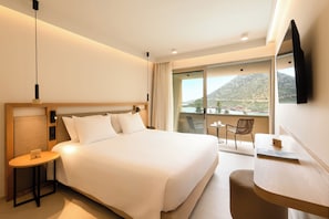 A hotel room with a large bed, a desk, a chair, a small table, a TV, and a view of a mountain.