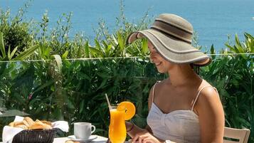 Uma pessoa almoçando em mesa ao ar livre com vista para o mar, uma cesta de pão, um croissant, um copo de suco de laranja e uma xícara.