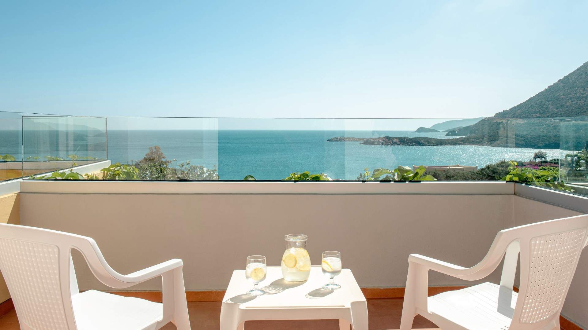 A balcony with a clear view of the sea and a distant island, two white chairs, and a table with a pitcher and glasses.