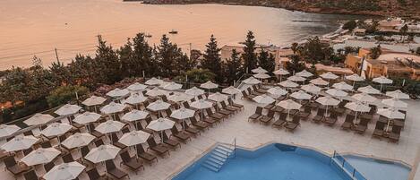 A poolside area with sun loungers and umbrellas, overlooking a body of water and a hilly landscape during sunset.