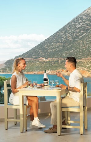 Two people dining at a table with a bottle of wine, glasses, and a view of a coastal landscape. - Mitsis Bali Paradise (Mylopotamos)