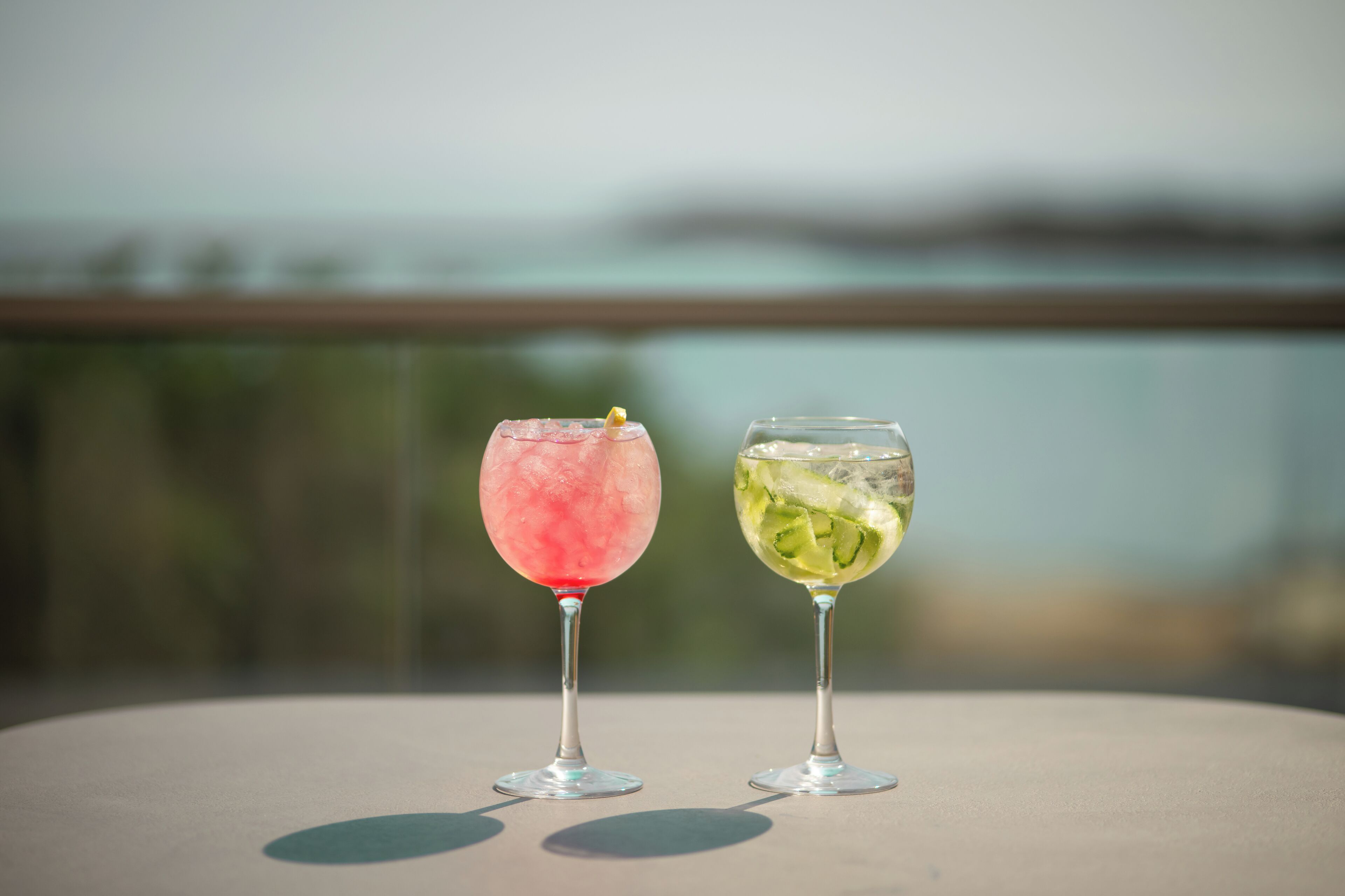 Two cocktails on a table with a blurred outdoor view in the background.