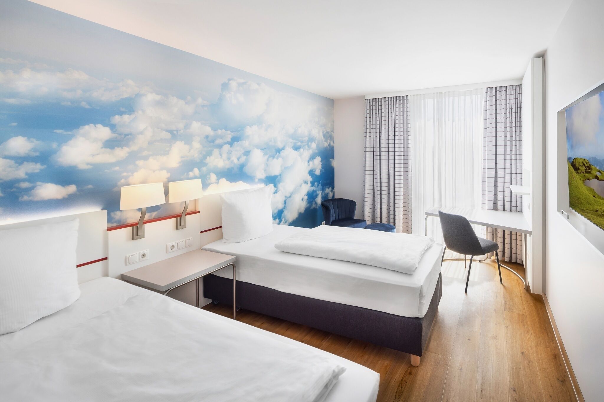 Una habitación de hotel con cama, escritorio, silla y un mural de nubes en la pared.