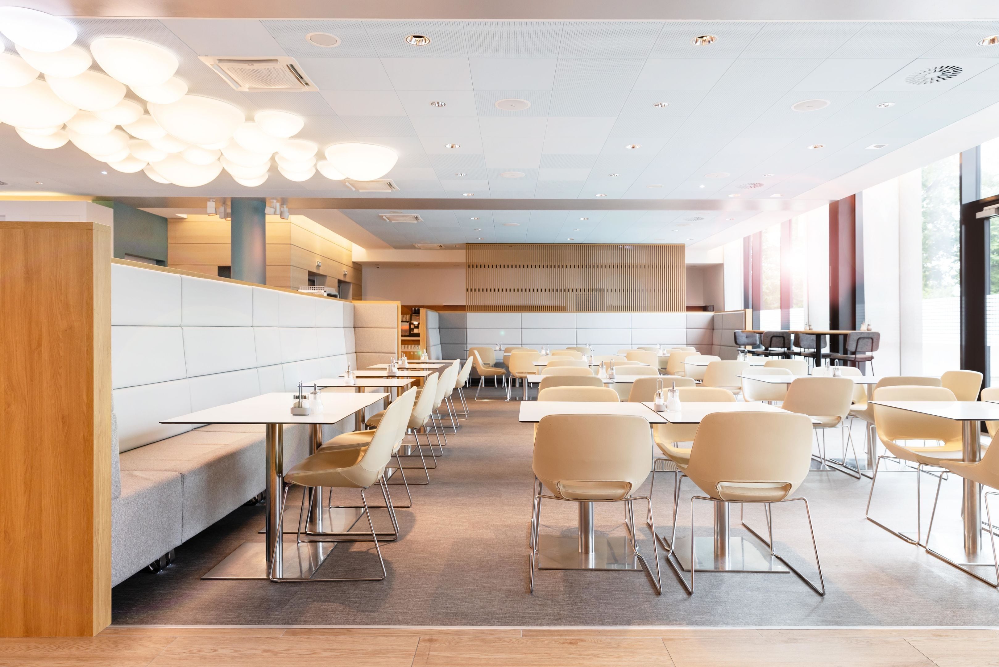A modern restaurant with long communal tables and booths, a wooden accent wall, and a ceiling with recessed lighting.