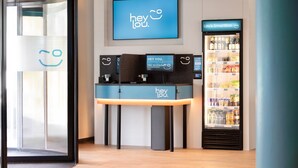 Un área de recepción con una pantalla digital mostrando “hey lou”, un mostrador de recepción y un refrigerador lleno de bebidas.
