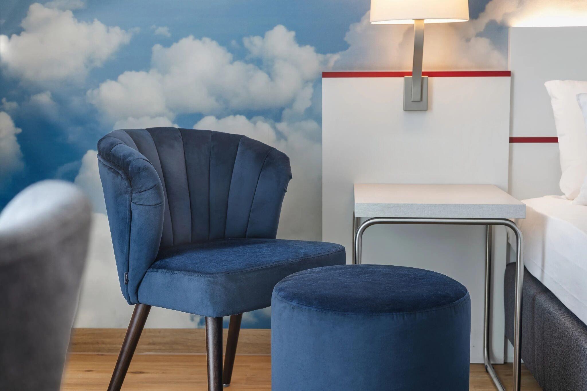A room with a blue chair, a small ottoman, a white bedside table, and a wall mural of clouds.