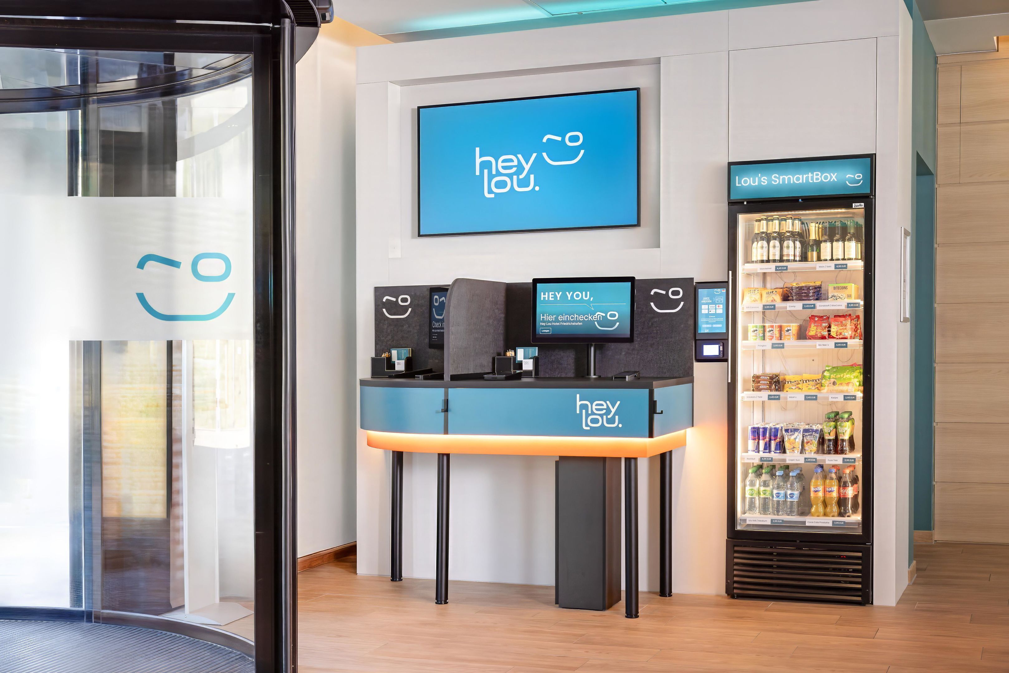 Un espace d’accueil doté d’un écran numérique affichant le message « hey lou » et d’un distributeur automatique portant l’inscription « Lou’s SmartBox ».