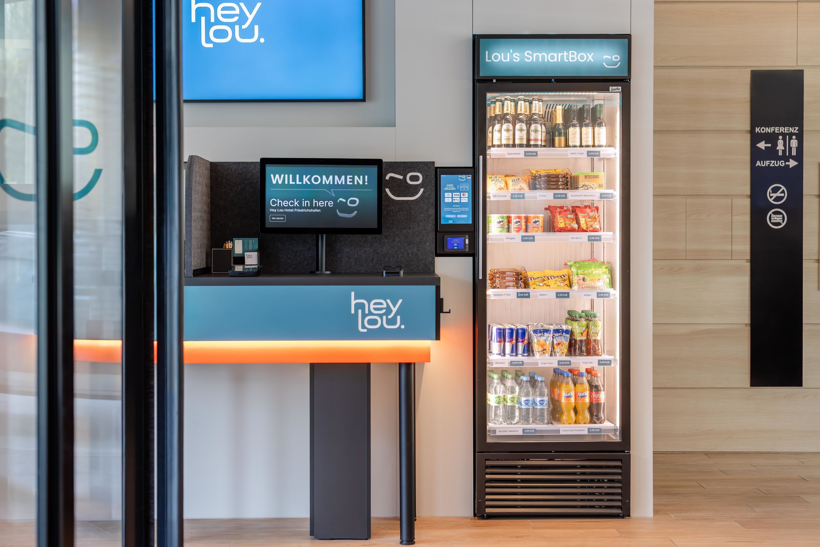 Ein Empfangsbereich mit einem digitalen Check-in Kiosk, einem mit Getränken gefüllten Lou's SmartBox Kühlschrank und Hinweisschildern für Konferenzräume und Ausgang.