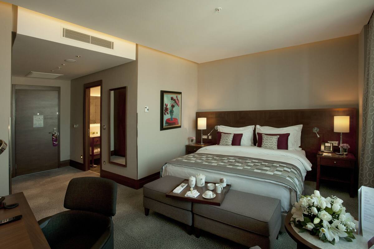 A hotel room with a large bed, a desk with a chair, a bedside table with a lamp, and a small table with a tray of breakfast items.