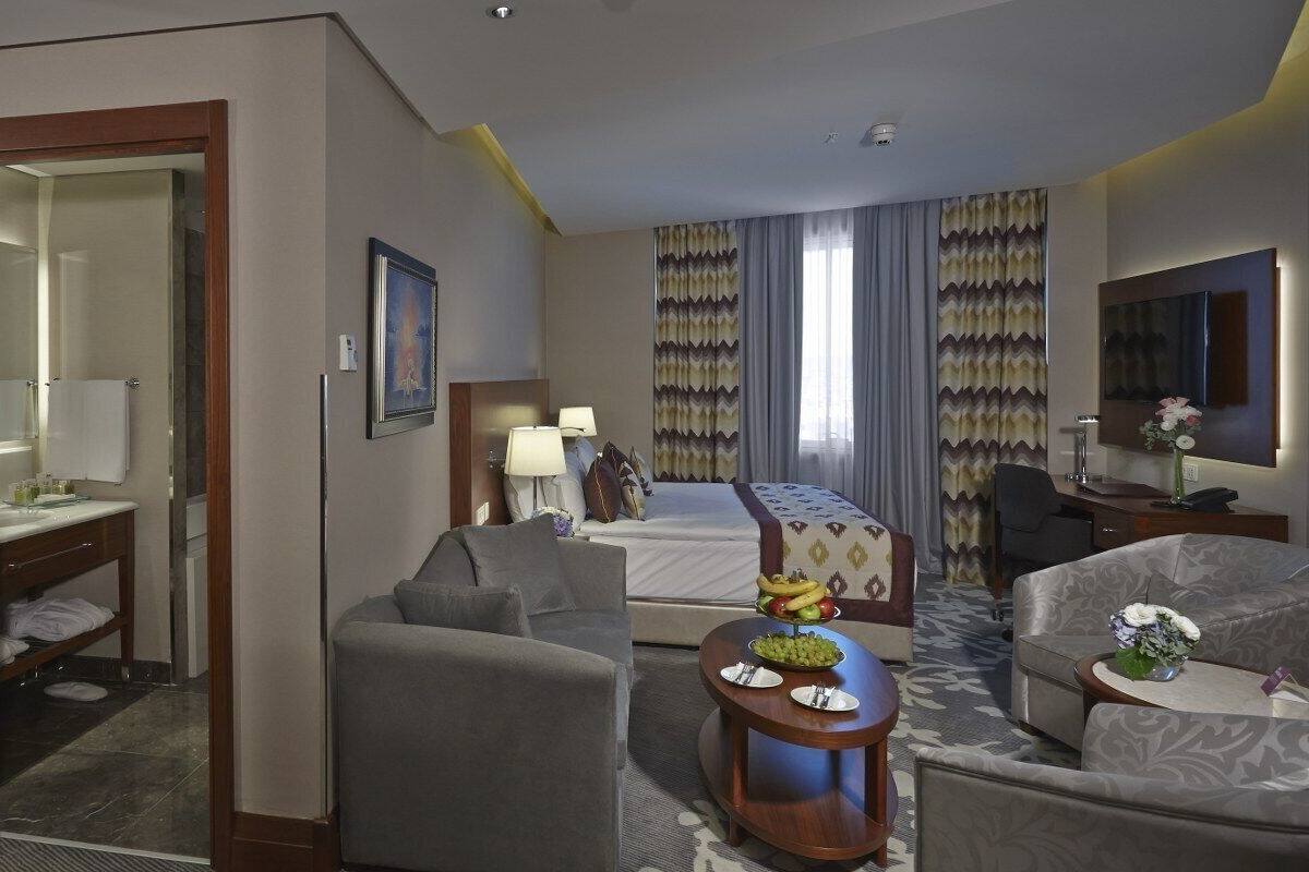 A hotel room with a bed, a desk with a chair, a sofa, a small table with fruit, and a TV.